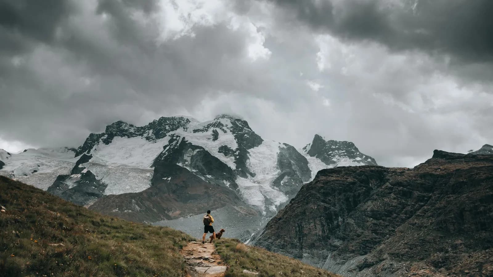 A hiker on a rugged mountain trail under dramatic clouds.